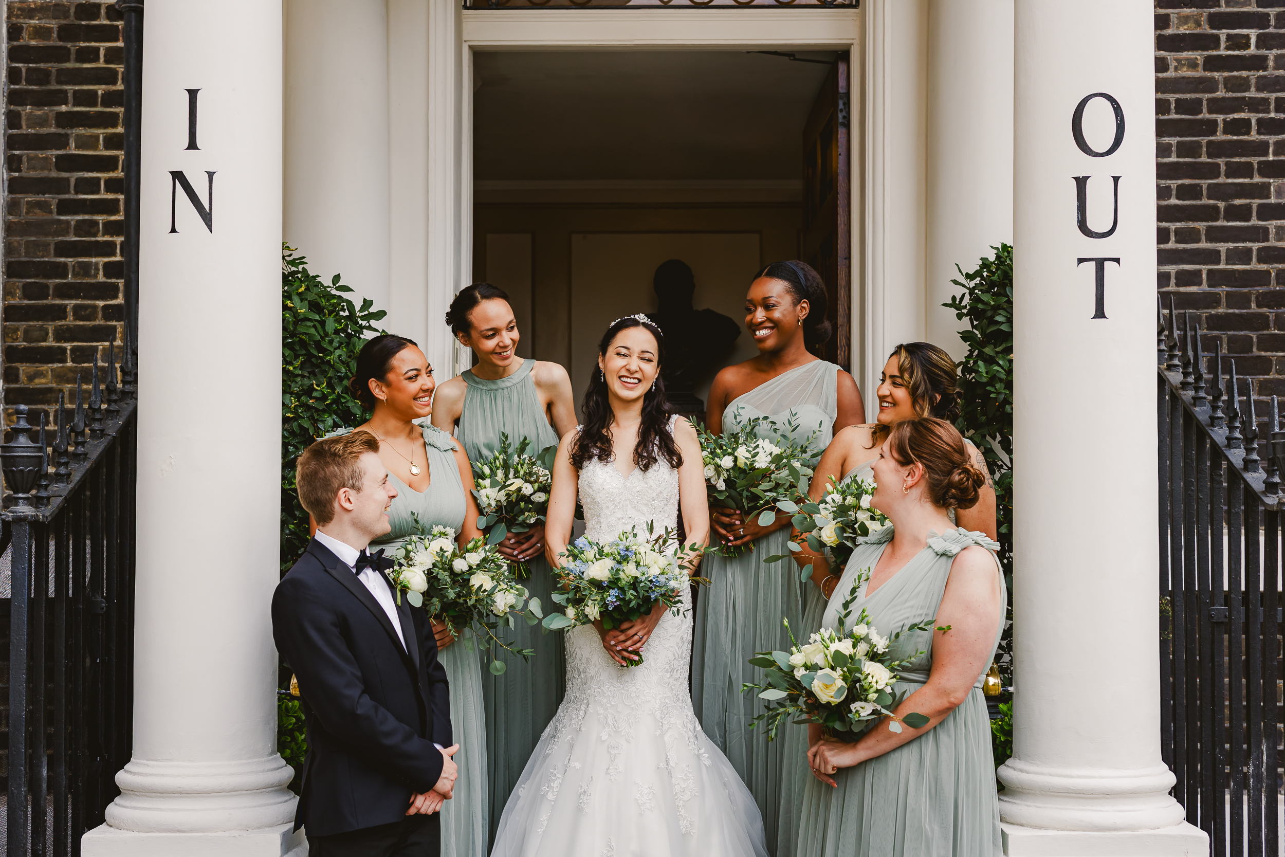 Bride and Bridesmaids on Steps of The In and Out Club – Wedding Party Portrait 1 Bride being gently helped downstairs inside The In and Out Club during wedding preparations