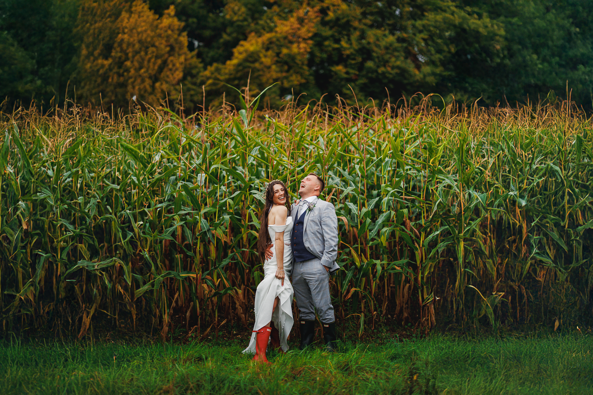 Oakley Hall Hotel Wedding: Alex and Matt’s Elegant Boho-Chic Wedding 3 Bride and groom laughing together in front of cornfields at Oakley Hall Hotel, bride playfully showing off her wellies.