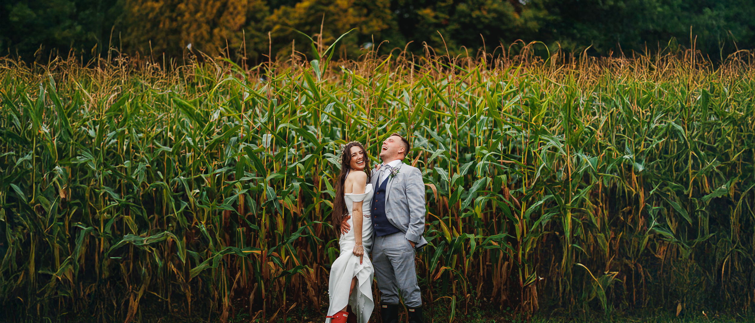 Bride and groom laughing together in front of cornfields at Oakley Hall Hotel, bride playfully showing off her wellies.